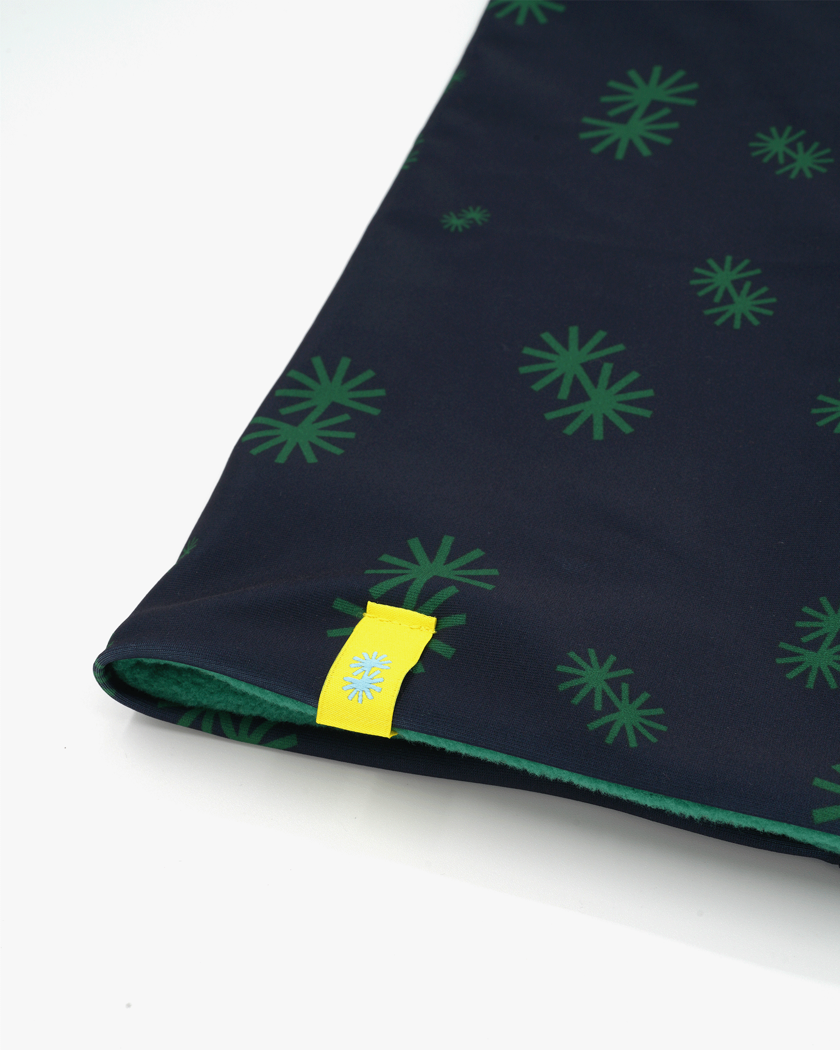 Close-up view of a double layer neck warmer with green snowflake on dark blue background geometric printed lycra, green fleece lining and yellow CTW logo woven label