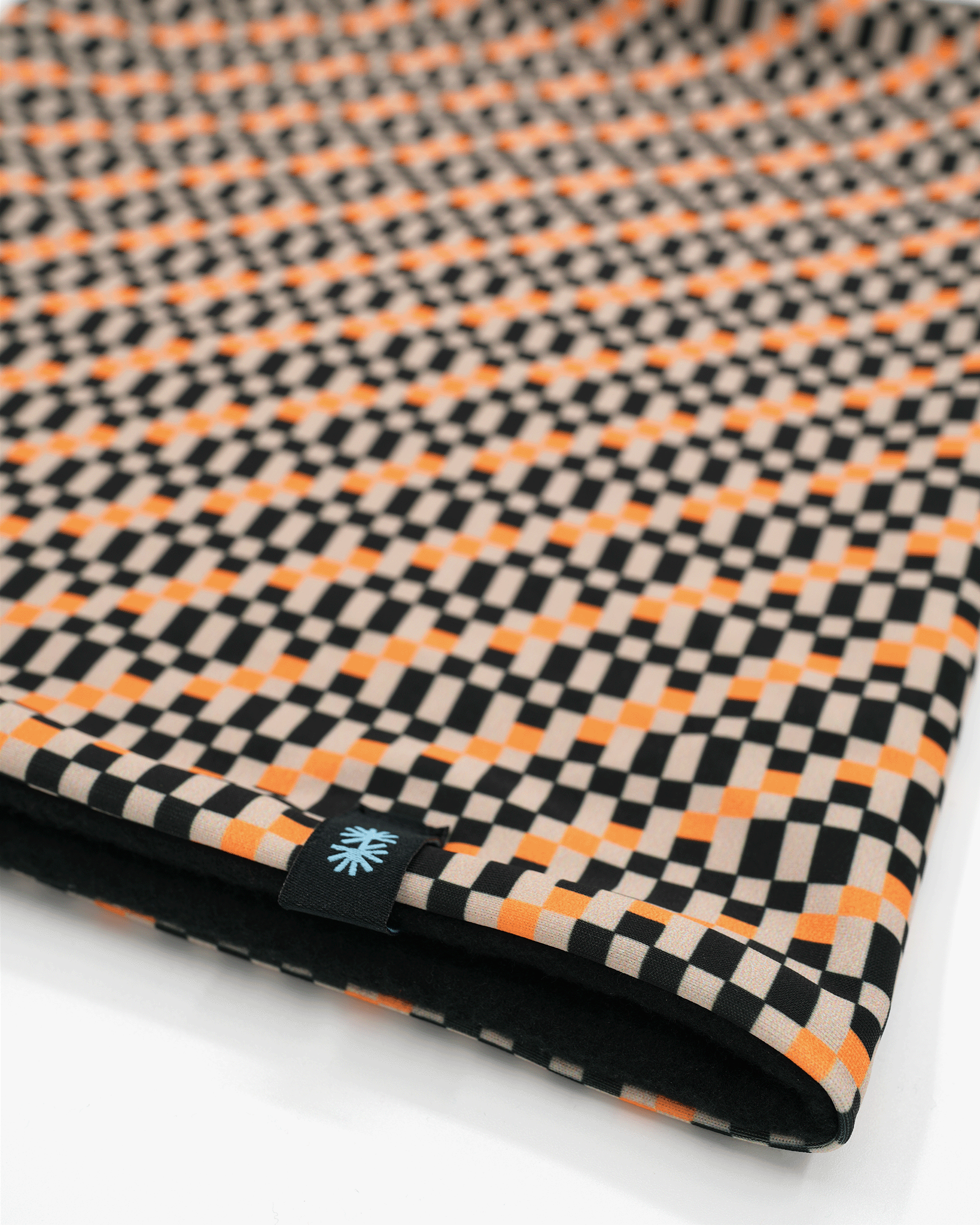 Close-up view of a double layer neck warmer with a black, orange and beige checkered printed lycra, black fleece lining and black CTW logo woven label