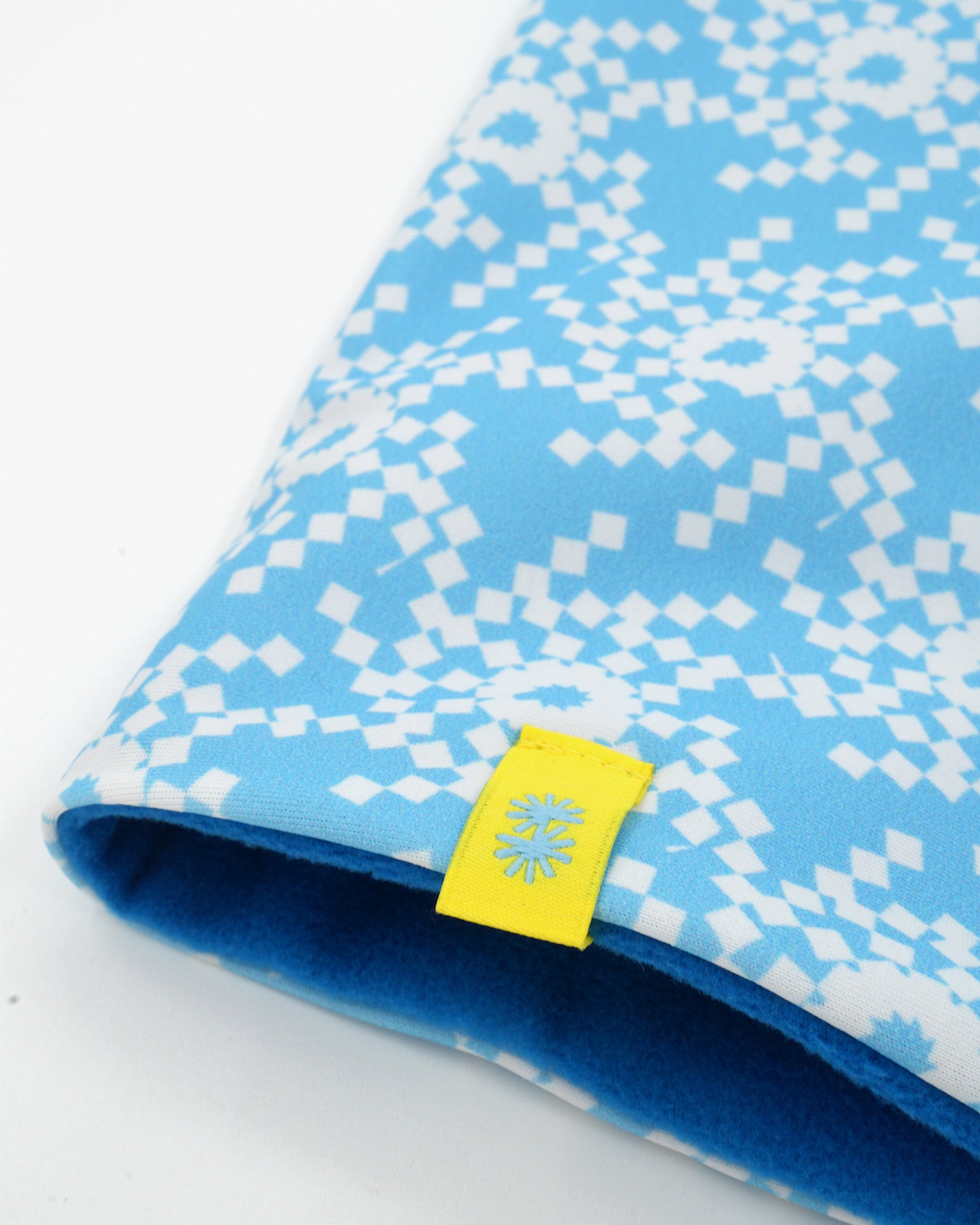 A close-up of a double layer neck warmer with blue and white geometric snowflake printed lycra, blue fleece lining and yellow CTW logo woven label