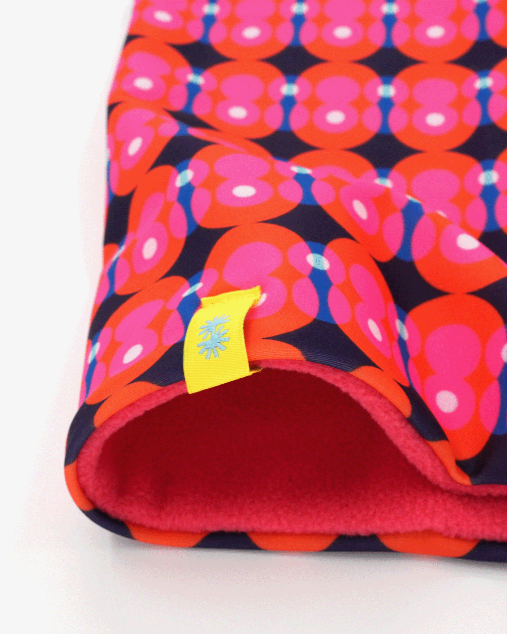 A close-up view of a double layer neck warmer with a bright pink and red circles on dark blue background geometric printed lycra, pink fleece lining and yellow CTW logo woven label